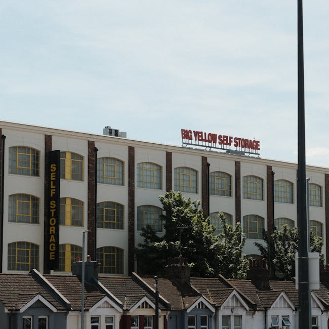 Exterior view of a multi-storey commercial building with a large sign reading 'BIG YELLOW SELF STORAGE' on the rooftop and another vertical sign on the side also indicating 'SELF STORAGE' in bold yellow letters. The building features a white facade with brown brick accents, large arched yellow-framed windows, and surrounding greenery of trees partially obscuring the lower levels. Below the storage facility, there are small residential-style houses with pitched tiled roofs and chimneys. The scene is illuminated by natural daylight, with a lamppost partially visible on the right side of the image, indicating an urban environment. This setting reflects typical locations involved in home relocation and furniture transport logistics, highlighting the connection between self-storage units and professional removal services, as operated by Removal Companies Highbury.