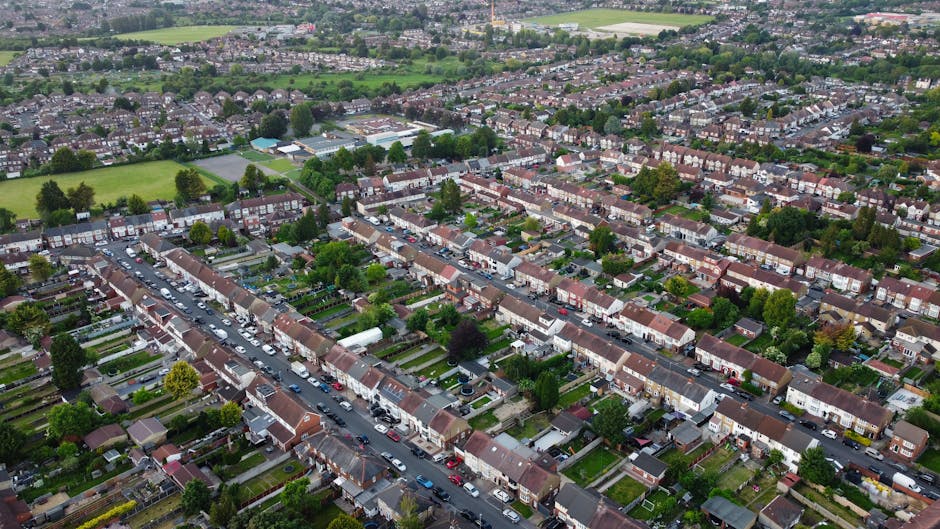Aerial view of a residential neighbourhood showing rows of terraced houses with pitched roofs, some with front gardens and driveways, lined along a main street filled with parked cars. Behind the houses, there are several parks and open green spaces, with trees and sports fields visible. The area is densely populated, with houses extending into the distance, surrounded by greenery and suburban development. This image captures the typical layout of a UK suburb, illustrating a quiet, well-organized community suitable for home relocations and household moves, which Removal Companies Highbury facilitates through their expert removals and packing services.