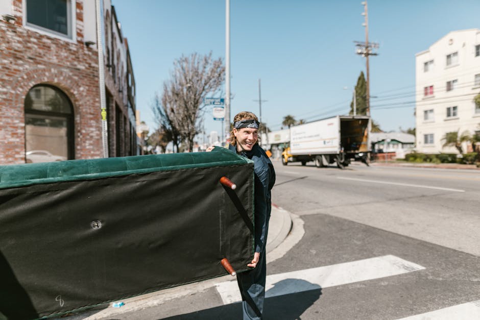 A person wearing a black jacket and a patterned headband is carrying a large, rectangular mattress wrapped in a dark protective cover with orange handles, during a home relocation process outside a brick building on a sunny day. The individual is smiling as they load the mattress onto a moving van parked on the street. The scene shows the city environment with a pedestrian crossing, a white delivery truck in motion, and residential buildings in the background. The mattress appears to be part of the furniture transport in a professional move coordinated by Removal Companies Highbury, emphasizing the packing and loading process involved in Highbury house removals, with the furniture being carefully prepared for transport at the doorway or pavement area.