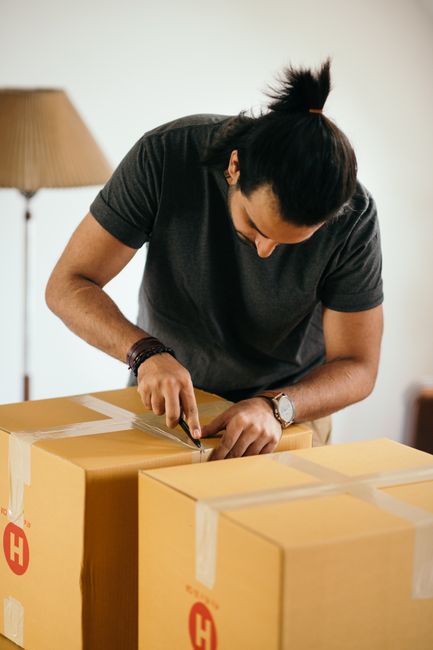 A person with dark hair tied back, wearing a dark grey t-shirt and a silver wristwatch, is seen inside a home, carefully sealing a cardboard box with packing tape using a dispenser. The individual is leaning over a wooden table or countertop, which holds multiple similarly packed boxes, some of which have printed labels or markings. The background features a warm-toned lamp on a side table, indicating a domestic setting. The scene appears to be part of a home relocation or furniture transport process overseen by Removal Companies Highbury, with attention to proper packing techniques for moving household items securely and efficiently. The lighting in the room is soft and natural, highlighting the focus on packing and consolidation of boxes in preparation for transportation or scanning for a home removal service.