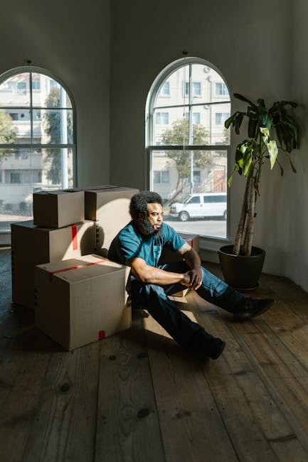 A man with curly dark hair and medium skin tone, wearing a blue T-shirt and jeans, sits on the wooden floor of a room near a large arched window. He appears to be taking a break during a home relocation process, with several packed cardboard boxes stacked behind him, some sealed with red tape. Natural daylight streams through the window, illuminating the space and revealing a view of parked cars and nearby buildings outside. A large potted plant with green leaves is positioned near the window, adding to the room's interior decor. The setting suggests an environment in the midst of moving, with the boxes ready for transport, and [COMPANY_NAME] likely involved in the packing or loading process as part of their removals services offered in Highbury. The scene emphasizes the preparation and logistics involved in furniture transport and house removals, with a focus on careful packing, staging, and the moving process within a residential property.