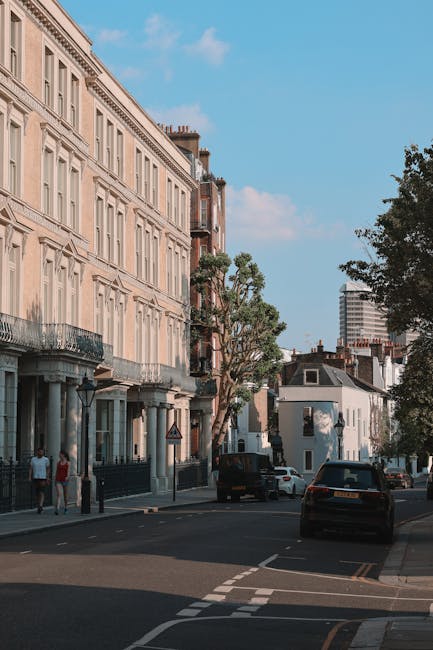 A view of a residential street in Highbury, showing a row of light-colored, multi-storey houses with ornate architectural details and balconies along the left side. The street is lined with parked cars, including a black vehicle near the foreground, and features a pavement with pedestrians walking. Several mature trees with green foliage are visible, providing shade along the sidewalk. In the background, modern high-rise buildings can be seen against a bright, blue sky with scattered clouds. The scene depicts an urban environment suitable for local house removals or furniture transport, with no active loading or moving process visible in the image.