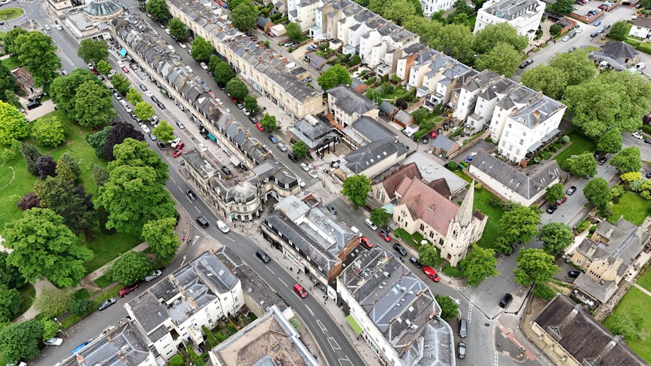 An aerial view of a residential street in Highbury, showing a mixture of terraced houses, church buildings, and surrounding green spaces with mature trees. The street is lined with parked cars and features a loading area near the curb, where a van from Removal Companies Highbury is actively being loaded with furniture and packed boxes covered in cardboard and plastic wrapping. The scene captures the logistics of a home relocation, with moving trolleys and securing straps visible as workers carefully handle household items, including sofas and small appliances, in the process of packing and transportation. The lush greenery and well-maintained properties highlight the typical environment for house removals in the area, with bright, natural daylight illuminating the scene, supporting professional furniture transport and moving services within Highbury.