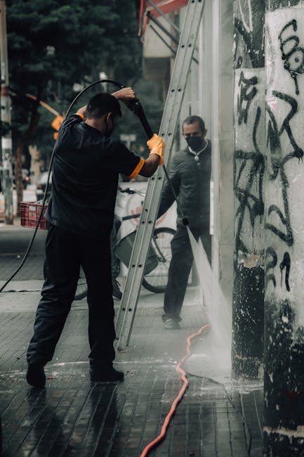 A worker from Removal Companies Highbury is performing a street cleaning task during a home relocation process, using a pressure washer to clean the pavement next to a graffiti-covered wall in an urban environment. The worker, dressed in dark clothing with orange gloves, stands on a wet, tiled sidewalk while holding a high-pressure hose, directing water towards the ground. Nearby, an aluminum ladder is propped against the wall, possibly used earlier for accessing higher areas. Behind the worker, an onlooker dressed in casual attire and a face mask observes the scene, standing near a doorway adjacent to the graffiti. The background shows blurred street elements and trees, indicating an outdoor setting with natural lighting. This scene reflects the cleanup and logistical aspects involved in house removals and urban furniture transport conducted by professionals such as those at Removal Companies Highbury, supporting a comprehensive house moving service.