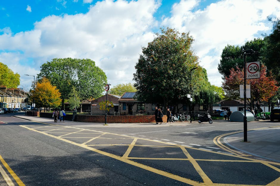 A street scene in a residential area showing a small brick building surrounded by large trees with green, yellow, and orange leaves, indicating autumn. The street has yellow box junction markings and a double yellow line on the curb, with a 20 mph speed zone sign visible. Several pedestrians and cyclists are present, with some waiting at the corner and others walking along the pavement. A black lamppost is positioned near the sidewalk. The sky is partly cloudy with patches of blue, and the overall scene suggests a calm local neighbourhood environment, potentially used for home relocations or community access, relevant to highbury removals and house moving services.