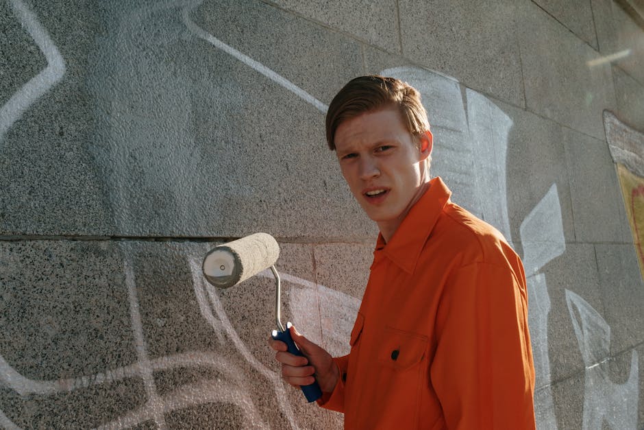 A worker from Removal Companies Highbury is performing a street cleaning task during a home relocation process, using a pressure washer to clean the pavement next to a graffiti-covered wall in an urban environment. The worker, dressed in dark clothing with orange gloves, stands on a wet, tiled sidewalk while holding a high-pressure hose, directing water towards the ground. Nearby, an aluminum ladder is propped against the wall, possibly used earlier for accessing higher areas. Behind the worker, an onlooker dressed in casual attire and a face mask observes the scene, standing near a doorway adjacent to the graffiti. The background shows blurred street elements and trees, indicating an outdoor setting with natural lighting. This scene reflects the cleanup and logistical aspects involved in house removals and urban furniture transport conducted by professionals such as those at Removal Companies Highbury, supporting a comprehensive house moving service.