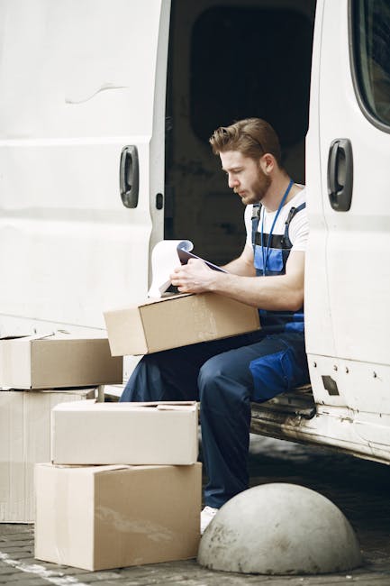 A young male removal worker with light skin, short brown hair, and a beard, wearing a striped t-shirt and blue overalls, is seated on the edge of a white van with sliding door open. He is holding a clipboard and appears to be reviewing a sheet of paper. In front of him on the ground are three cardboard boxes, some open and partially filled, with additional boxes nearby. The van is parked in an outdoor area with a concrete bollard and a paved surface visible. The interior of the van, which is used for home relocation and furniture transport, can be seen in the background. This scene depicts a packing and loading process relevant to professional removals services, with the worker organizing documentation during the loading process, supported by tools like boxes and the vehicle for efficient house removals.