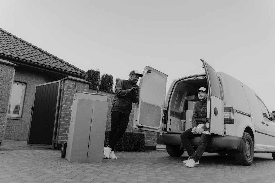 Two men engaged in a home relocation process as part of a furniture transport activity, with one standing outside near the open rear doors of a white van and the other seated on the van’s bumper. The man outside is dressed in a dark jacket and cap, holding a smartphone, while the man inside wears a hat and dark clothing, smiling at the camera. In front of the van, there are several large cardboard boxes, along with packing materials such as bubble wrap and kraft paper, indicating packing and moving preparations. The van is parked on a paved driveway adjacent to a residential building with a tiled roof and brick exterior, with a small garden and shrubbery visible in the background. The loading process appears to be ongoing, with the van's rear doors fully open, ready for the transportation of household furniture and boxes. The environment is well-lit with natural light, and the scene is captured in black and white, emphasizing the practical aspects of house removals and logistics facilitated by [COMPANY_NAME].
