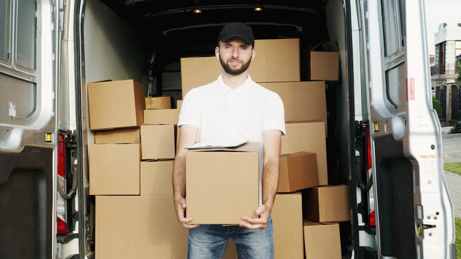 A young male removal worker with light skin, short brown hair, and a beard, wearing a striped t-shirt and blue overalls, is seated on the edge of a white van with sliding door open. He is holding a clipboard and appears to be reviewing a sheet of paper. In front of him on the ground are three cardboard boxes, some open and partially filled, with additional boxes nearby. The van is parked in an outdoor area with a concrete bollard and a paved surface visible. The interior of the van, which is used for home relocation and furniture transport, can be seen in the background. This scene depicts a packing and loading process relevant to professional removals services, with the worker organizing documentation during the loading process, supported by tools like boxes and the vehicle for efficient house removals.
