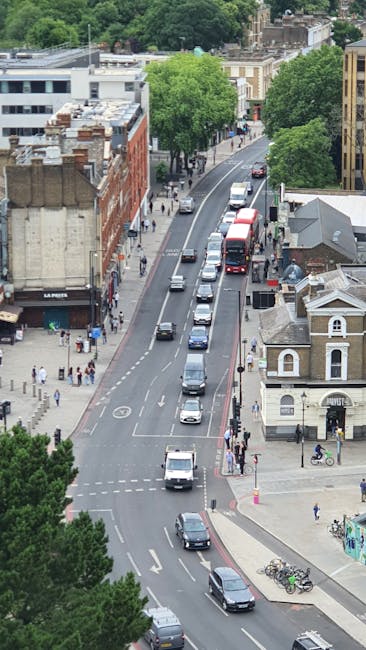 An aerial view of a busy urban street scene with a row of parked vehicles, including a white van and several small cars, lined along the curb on the right side. Moving vehicles, such as a red double-decker bus and a dark-coloured car, are driving along the road. The scene captures the typical environment of a home relocation process, with trees lining the street and a mix of commercial and residential buildings, including a brick building with a sign, small shops, and houses. Pedestrians are walking on the sidewalks, some near the buildings and others crossing the street, reflecting an active neighbourhood area. In the background, lush green trees provide a natural contrast to the urban setting, and the overall lighting suggests daytime conditions. This image illustrates the urban logistics involved in furniture transport and packing during home moves, aligned with removal services offered by companies like Removal Companies Highbury.