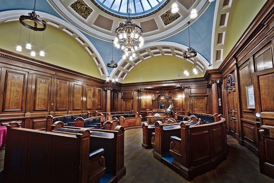 Inside a historic, ornate chamber with polished dark wood paneling and curved wooden benches arranged in a semicircular formation. The ceiling features a decorative, domed design painted in soft shades of blue and cream, with a large glass skylight allowing natural light to illuminate the room. Elegant chandeliers and vintage-style pendant lighting hang from the ceiling, enhancing the classical ambiance. The walls display plaques or inscriptions, possibly memorials or honorary listings, integrated into the wooden paneling. The room appears to be used for official or ceremonial purposes, with a central elevated area at the front. Although no furniture or moving equipment is visible, the detailed interior suggests a setting where careful furniture transport and handling might occur if relocation services, such as those by Removal Companies Highbury, were involved in moving items during a house or office relocation process.