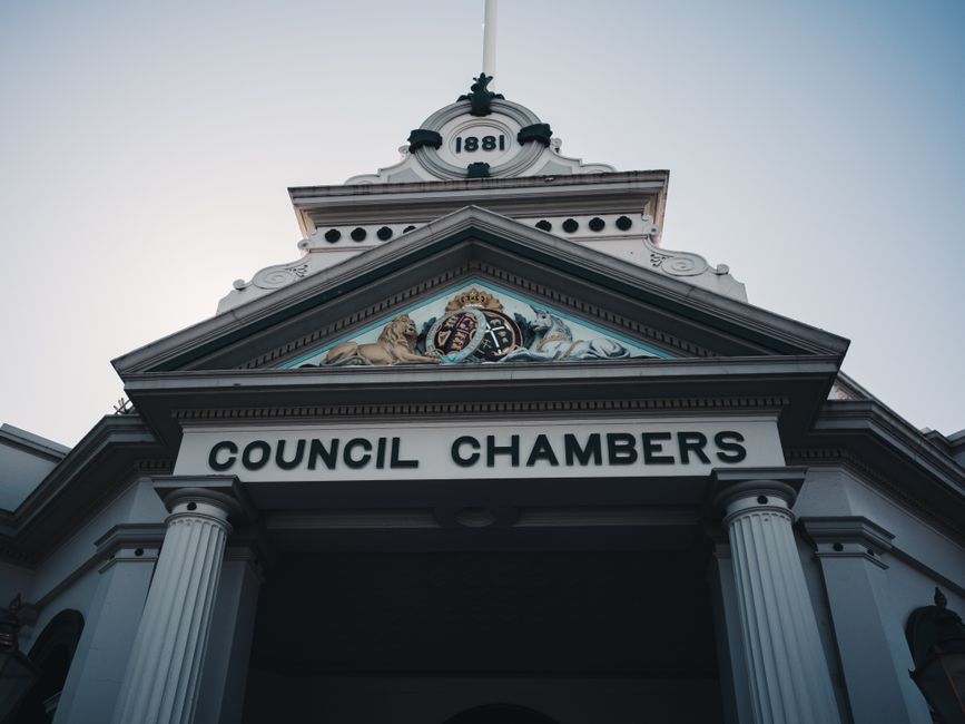 A low-angle photograph of a historic council chambers building, showing its classical architectural features including white columns supporting a pediment with an ornate crest and the year '1881' displayed at the top. The building's façade is painted in light colours with decorative details, and the sky in the background is clear, providing natural daylight that illuminates the structure. The imagery suggests a civic or government building in Highbury, associated with local council activities relevant to house removals and relocation services offered by Removal Companies Highbury, as indicated by the website page titled 'What to know about Islington Council permits for Highbury removals.' The scene captures elements related to the logistical aspects of home relocation, such as permits, planning, and civic regulations, subtly supported by the formal appearance of the council chambers.